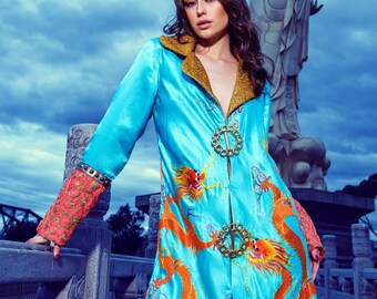 May include: A woman wearing a turquoise blue satin jacket with gold trim and embroidered orange dragons. The jacket has a gold chain zipper and decorative gold buttons.