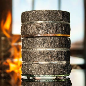 May include: A decorative, stacked object made of dark grey, textured stone segments, separated by clear and coloured glass rings. The object is reflected on a glossy surface, with a blurred background of a window and a warm fire.