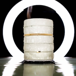 Könnte beinhalten: Ein geschichtetes, zylindrisches Objekt mit einem goldenen Band, das einen Rauchwölkchen vor einem hellen weißen Ring auf schwarzem Hintergrund abgibt. Das Objekt scheint aus gestapelten, durchscheinenden Blöcken zu bestehen.