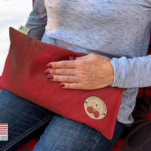 May include: Red cherry pit pillow with a brown label that says "Cherry Pit Pillow" with a heart and two cherries. The pillow is being held by a person wearing blue jeans and a gray shirt.