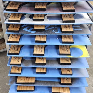 May include: Wooden plaques with a dark border and light wood grain are arranged on shelves. The plaques have a unique shape and feature the text "March 2025" and "February 25". The shelves are blue and the background is gray.