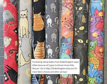 May include: Seven long, cylindrical catnip kicker toys featuring various cat-themed fabric patterns in red, grey, black, and multicoloured prints. A white rectangular sign with black text is centred over the toys, detailing a charitable donation.