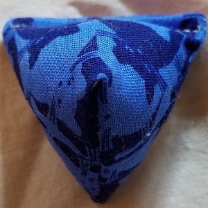 May include: A small, triangular fabric pouch featuring a two-tone blue pattern. The textile has a light blue base color with a darker navy blue abstract print. The object is stitched at the corners, creating a three-dimensional pyramid shape.