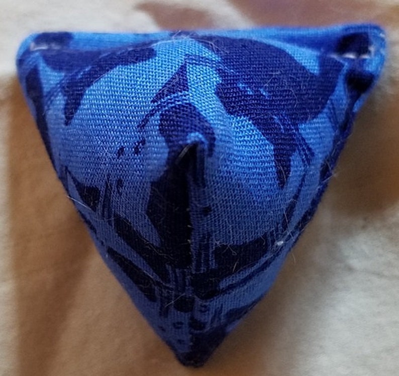May include: A small, triangular fabric pouch featuring a two-tone blue pattern. The textile has a light blue base color with a darker navy blue abstract print. The object is stitched at the corners, creating a three-dimensional pyramid shape.
