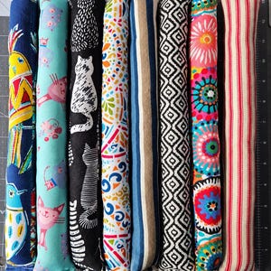 May include: Eight long, cylindrical fabric tubes arranged side-by-side. Patterns include a yellow bird, light blue cats, black cats, colorful geometric shapes, blue and tan stripes, black and white diamonds, vibrant floral mandalas, and red stripes.