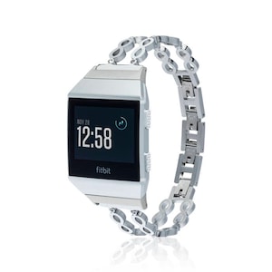 Puede incluir: Un reloj inteligente Fitbit plateado con una banda de metal plateado. La esfera del reloj muestra la fecha "NOV 28" y la hora "12:58".