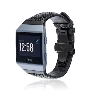 Puede incluir: Una correa de reloj de cuero negro con un patrón texturizado y una hebilla de acero inoxidable. La correa está unida a un reloj inteligente Fitbit plateado con una pantalla negra que muestra la hora "12:58" y la fecha "NOV 28".