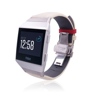 Puede incluir: Un reloj inteligente Fitbit plateado con una correa de cuero blanca y un acento rojo en la hebilla. La esfera del reloj muestra la fecha, la hora y un indicador de progreso.