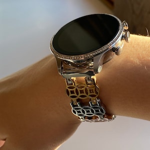 May include: A silver smartwatch with a black face and a gold and silver patterned metal band.