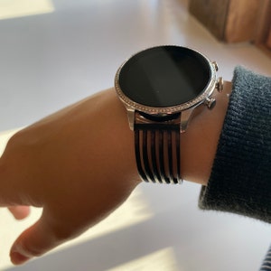 May include: A silver smartwatch with a black band and a diamond-encrusted bezel. The watch face is black and digital.