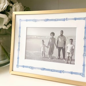May include: A gold picture frame with a white mat and a blue bamboo border. The frame contains a black and white photo of a family of four on a beach.