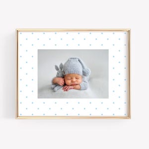 May include: A framed photograph featuring a newborn baby wearing a light blue knit cap and matching outfit, cradling a small stuffed bunny. The frame is gold with a white border adorned with light blue polka dots. The baby is sleeping peacefully.
