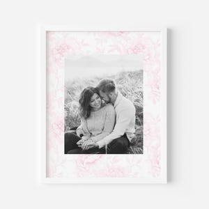 May include: Black and white photo of a couple sitting in a field of tall grass. The woman is leaning against the man's shoulder and they are both smiling. The photo is framed with a pink floral border.
