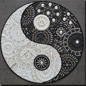 May include: A black and white yin yang symbol made of beads, with intricate patterns and designs. The symbol is on a dark gray background.
