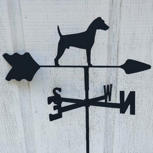 May include: Black metal weather vane featuring a silhouette of a dog with a pointed tail, facing right. The vane has a directional arrow and compass points.