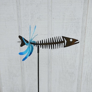 May include: A metal fish skeleton wind spinner with blue and green fins. The spinner is mounted on a black metal rod.