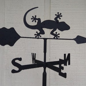 May include: Black metal weather vane featuring a silhouette of a gecko with a compass rose and directional arrows. The arrows point north, south, east, and west.