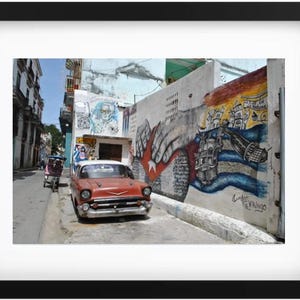 Op de afbeelding: Een rode vintage auto geparkeerd in een smalle straat in een stad. De auto staat voor een gebouw met een kleurrijke muurschildering met een hand die reikt en een Cubaanse vlag. De muurschildering bevat ook de tekst "Cuba"