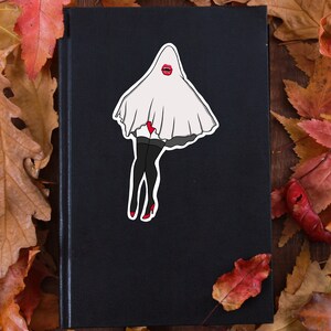 Puede incluir: Una pegatina de fantasma blanco con labios rojos y medias negras sobre un libro negro con fondo de hojas de otoño.