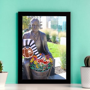 Op de afbeelding: Een bronzen beeld van een man die op een bank zit met een zwart-wit gestreept been en een rode schoen die uit een emmer met bloemen steekt. Het beeld staat voor een gebouw met grote ramen.