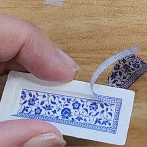 May include: A rectangular sticker with a blue floral pattern and a decorative border is being peeled away from a white backing. The design features stylized flowers and leaves. The sticker is partially peeled, revealing the adhesive side.