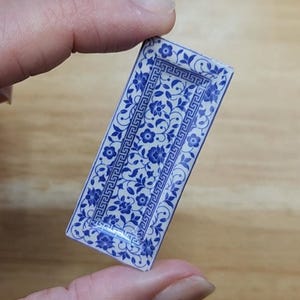 May include: A small, rectangular blue and white decorative tray with a floral pattern. The tray is held between fingers and features a detailed design with flowers and geometric borders, suitable for miniature displays or dollhouses.