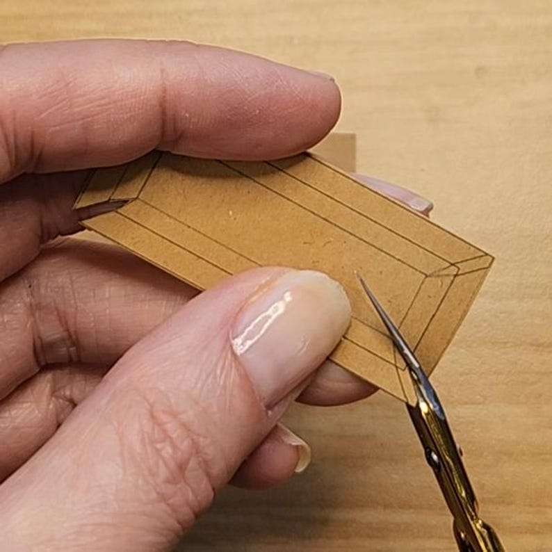 May include: A person's hand holding a small, brown paper box with black lines, being cut with small gold scissors. The box appears to be a miniature craft project, possibly for a dollhouse or other small-scale creation.