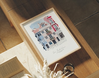 May include: A framed print featuring a watercolor illustration of colorful buildings with red, blue, and pink facades. The print is in a light wood frame and has the text "Our First Home" at the bottom. The print is displayed on a wooden surface.