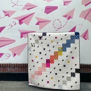 Puede incluir: Una colcha blanca con un patrón geométrico de triángulos negros y cuadrados coloridos. La colcha está hecha de diferentes telas con varios patrones y colores, incluyendo rosa, rojo, amarillo, azul y gris.