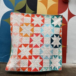 May include: A vibrant quilt featuring a star pattern in red, orange, and teal. The quilt is set against a wall with a rainbow mural. The design of the quilt showcases a repeating star pattern, adding a playful element to the scene.
