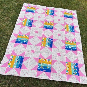 Puede incluir: Una colcha rosa y blanca con un patrón de estrellas. Las estrellas están hechas de diferentes telas con rayas azules, amarillas y naranjas.