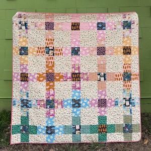 Puede incluir: Un edredón de patchwork colorido con un patrón de cuadrícula. El edredón presenta cuadrados de varios colores, incluyendo rosa, naranja, azul y verde, con patrones florales y geométricos. El edredón se muestra contra una pared verde.