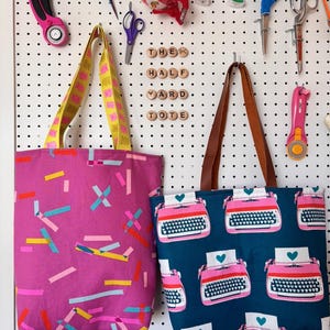 May include: Two tote bags hang on a white pegboard. One is magenta with colourful geometric shapes, the other is navy blue with a pink typewriter pattern. The text "THE HALF YARD TOTE" is spelled out with wooden letter beads.
