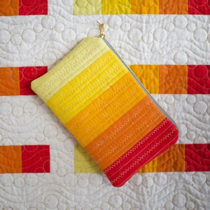 Puede incluir: Una bolsa con cremallera de rayas amarillas, naranjas y rojas con un tirador de cremallera dorado. La bolsa está sobre una tela acolchada blanca con un patrón de círculos.