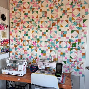 Peut inclure: Une courtepointe patchwork colorée avec des motifs d'étoiles et de carrés est accrochée au mur. Deux machines à coudre sont posées sur une table en bois, avec un tabouret bleu et une chaise blanche. La courtepointe présente des formes géométriques roses, bleues, vertes et oranges.