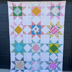 Może przedstawiać: Ręcznie robiona kołdra z wzorem gwiazdy. Kołdra ma białe tło z małymi różowymi kropkami. Każda gwiazda wykonana jest z kolorowych tkanin we wzory w odcieniach niebieskiego, zielonego, żółtego, różowego i czerwonego. Kołdra wisi na ciemnym tle.