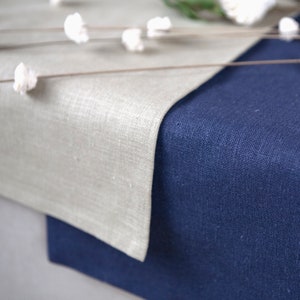 May include: Close-up of a linen table runner in two colors: a light beige and a deep navy blue. The fabric has a textured appearance. White floral accents are scattered across the top, adding a touch of elegance to the scene.
