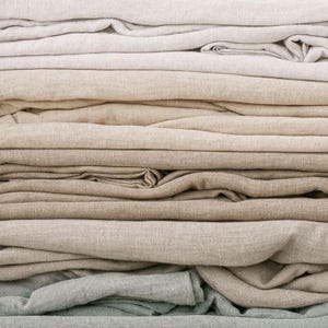 May include: A stack of folded fabric pieces in various neutral shades, including off-white, beige, tan, light brown, and soft sage green. The textiles feature a visible woven texture with natural creases and folds throughout the layers.