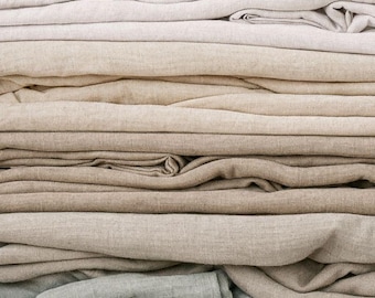 May include: A stack of folded fabric pieces in various neutral shades, including off-white, beige, tan, light brown, and soft sage green. The textiles feature a visible woven texture with natural creases and folds throughout the layers.