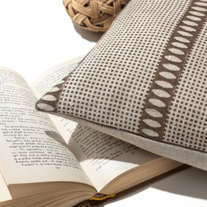 Puede incluir: Una almohada de tiro de color marr&oacute;n y blanco con un dise&ntilde;o geom&eacute;trico. La almohada est&aacute; hecha de una tela suave, similar al lino, y tiene un brillo sutil.