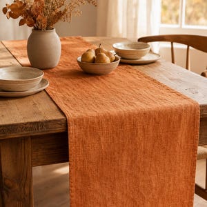 May include: A rectangular table runner in a solid burnt orange color with a visible woven texture. The runner is laid flat across the center of a wooden table, extending over the edge, featuring hemmed edges and a consistent fabric weave throughout.