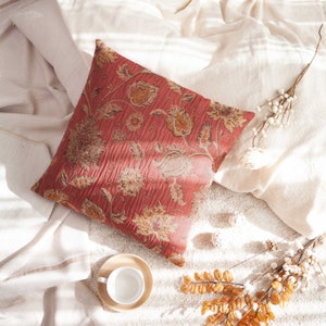 May include: A red floral patterned throw pillow with a quilted texture, resting on a white blanket with dried branches and a white ceramic cup on a wooden saucer.