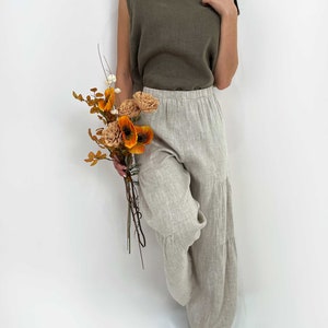 May include: A sleeveless olive green top paired with loose-fitting, tiered beige linen trousers. The outfit is complemented by a small bouquet of dried orange and brown flowers, featuring textured petals and thin stems held in a natural arrangement.