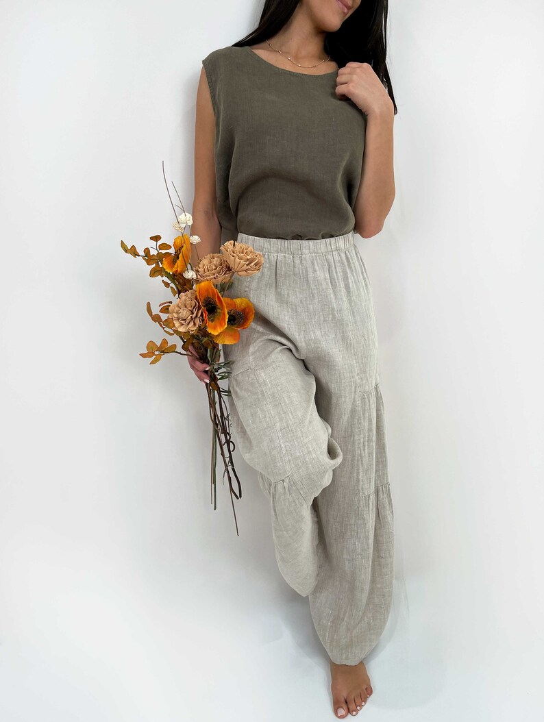 May include: A sleeveless olive green top paired with loose-fitting, tiered beige linen trousers. The outfit is complemented by a small bouquet of dried orange and brown flowers, featuring textured petals and thin stems held in a natural arrangement.