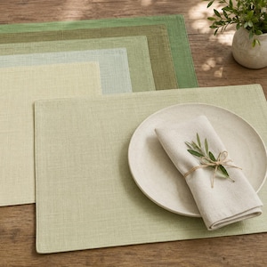 May include: A set of rectangular placemats in various shades of green and cream, featuring a woven texture. A round, speckled cream plate sits on the foreground placemat, topped with a folded cream napkin and a small green sprig tied with twine.
