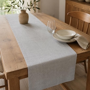 May include: A rectangular light gray table runner with a visible woven texture, laid flat across the center of a wooden dining table. The runner features clean, straight edges and a consistent neutral color throughout its entire length.