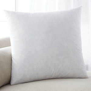 May include: A white, square pillow with a soft, smooth texture. The pillow is lying on a light beige surface.