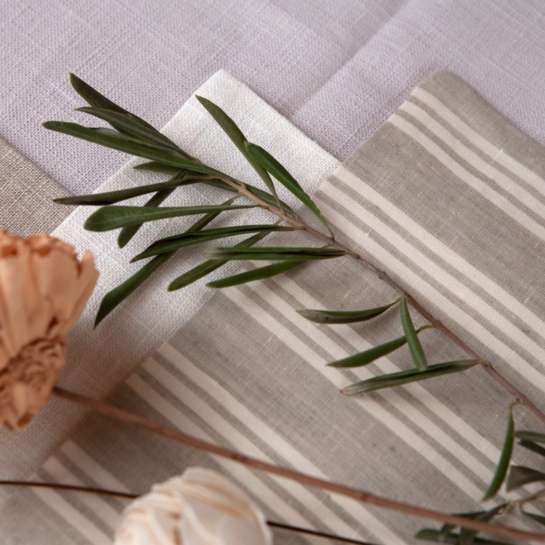 May include: A close-up of three different fabric swatches. The swatches are in shades of beige and white. The top swatch is a solid beige, the middle swatch is a solid white, and the bottom swatch is a striped beige and white fabric. A sprig of green leaves is placed on top of the swatches.