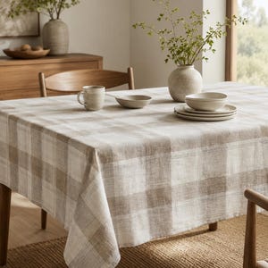 Puede incluir: Un mantel rectangular con un patrón de cuadros grandes en beige y blanco. La tela tiene una textura tejida visible y cuelga sobre los bordes de una mesa de comedor. La superficie sostiene una taza de cerámica, un cuenco y platos.