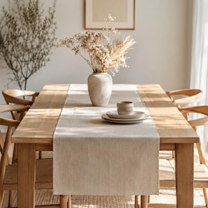May include: A light beige linen table runner centered on a rectangular wooden dining table. The table features a ceramic vase filled with dried flowers and pampas grass, alongside a stack of two ceramic plates and a small matching ceramic cup.